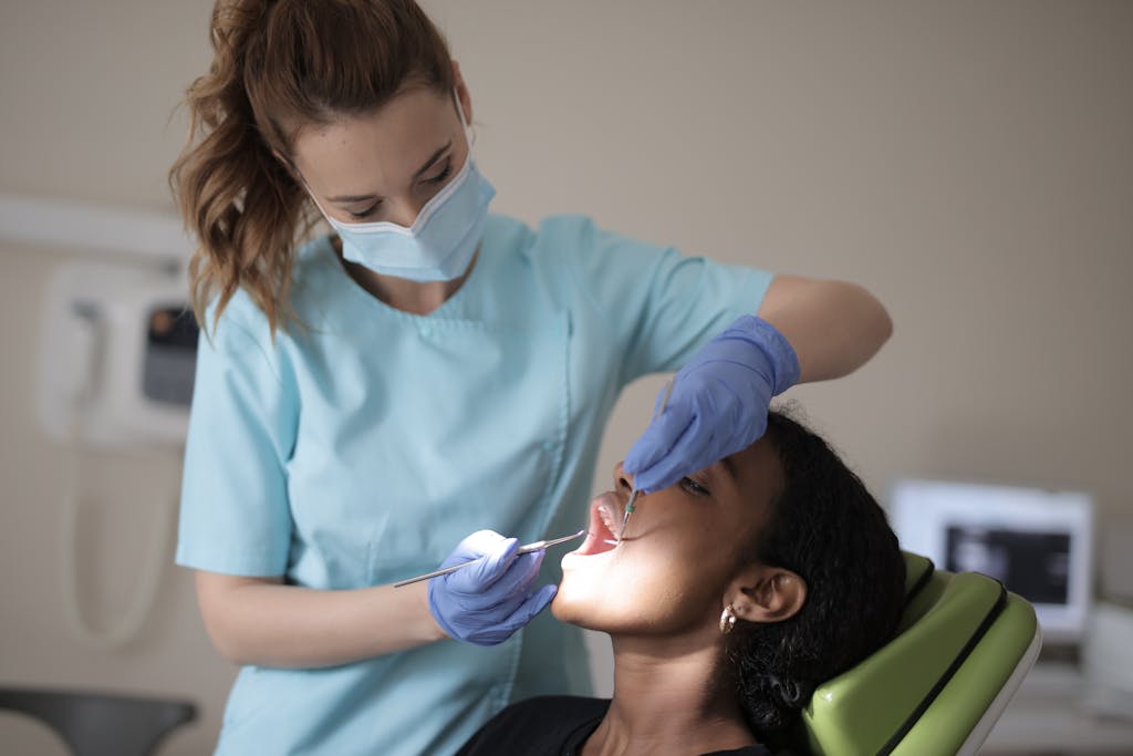 Dentist in blue scrubs and mask examines patient's teeth in clinic setting.
